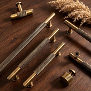 Puede incluir: Una variedad de tiradores y pomos de latón antiguo para armarios, dispuestos sobre una superficie de madera oscura. El herraje presenta un diseño acanalado y un acabado cálido y envejecido. También hay hierba seca decorativa.