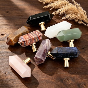 May include: A selection of gemstone cabinet knobs in diverse colours, such as black, white, green, purple, and pink. Each knob features a faceted, crystal-like form and is mounted on a gold-coloured base. The knobs are displayed on a dark wood surface.