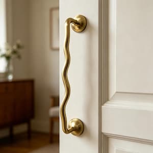 May include: A brass door pull with a wavy, organic design. The pull is mounted on a white door and has a curved shape. The door pull measures approximately 30 cm in length.