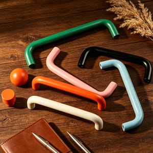 Peut inclure: Plusieurs poignées métalliques courbes de couleurs vives : vert, noir, bleu clair, rose, orange et crème. Une sphère et un cylindre orange sont également visibles. L'ensemble est disposé sur une surface en bois.