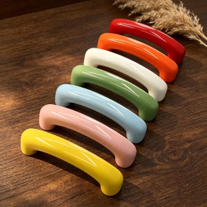 Puede incluir: Una colección de siete tiradores de armario coloridos en fila. Los tiradores son de color rojo, naranja, blanco, verde, azul claro, rosa y amarillo. Son de material brillante y están colocados sobre una superficie de madera.