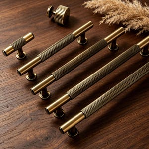 May include: A selection of antique brass cabinet hardware, including several long bar pulls, a small knob, and a T-shaped pull. The hardware features a ribbed centre section and a warm, aged finish, set against a dark wood surface.