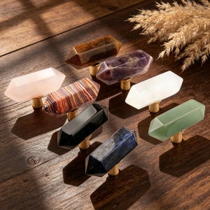 May include: A collection of crystal cabinet knobs in various colors, including pink, brown, black, and green. Each knob has a faceted, elongated shape and is mounted on a gold-toned base. The knobs are arranged on a wooden surface.