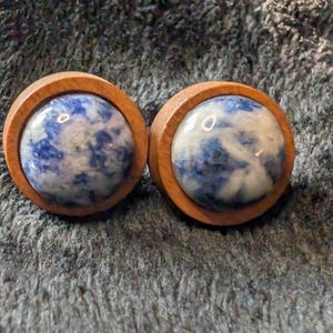 Puede incluir: Dos tapones para los oídos de madera con un centro redondo de piedra pulida azul y blanca. La madera tiene un tono marrón cálido y la piedra tiene un aspecto veteado. Los tapones están diseñados para perforaciones de orejas estiradas.