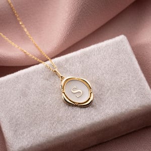 May include: A gold-colored necklace with a pendant featuring the letter "S" in a white oval shape. The pendant is set in a gold frame and hangs on a delicate gold chain. The necklace rests on a light gray velvet surface.