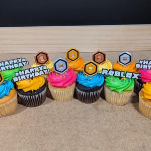 Op de afbeelding: Een verzameling van negen cupcakes versierd met kleurrijk glazuur en Roblox-thema toppers. De toppers omvatten gouden en zilveren munten, en de woorden "Happy Birthday!" en "Roblox".