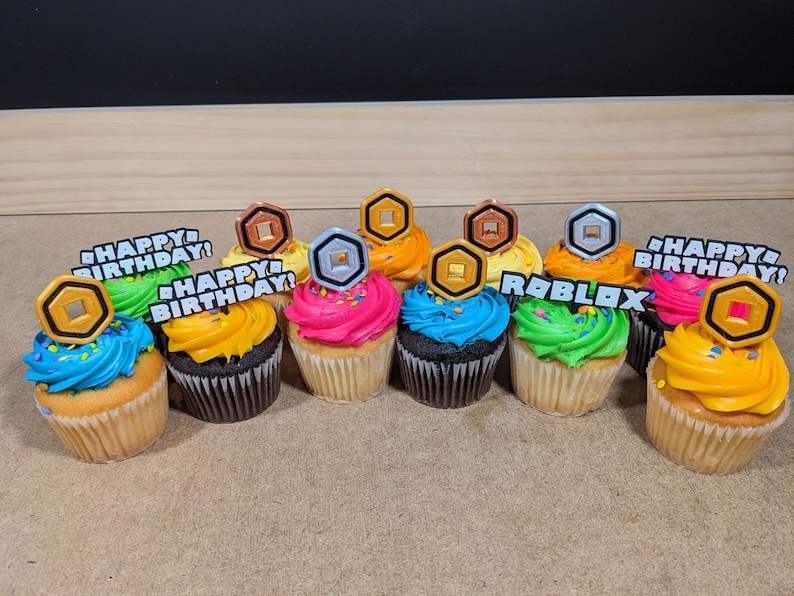 Op de afbeelding: Een verzameling van negen cupcakes versierd met kleurrijk glazuur en Roblox-thema toppers. De toppers omvatten gouden en zilveren munten, en de woorden "Happy Birthday!" en "Roblox".