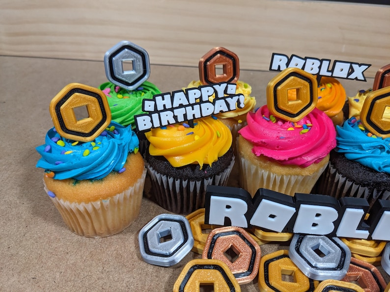 Op de afbeelding: Zes cupcakes versierd met kleurrijk glazuur en Roblox-thema toppers. De toppers omvatten gouden munten, een zilveren munt en het woord "Roblox". E&eacute;n cupcake heeft een topper met de tekst "Happy Birthday!"