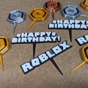 Op de afbeelding: Een set van 3D-geprinte taarttoppers met een Roblox-thema. De toppers bevatten een gouden munt, een zilveren munt en de woorden "Happy Birthday!" en "Roblox".