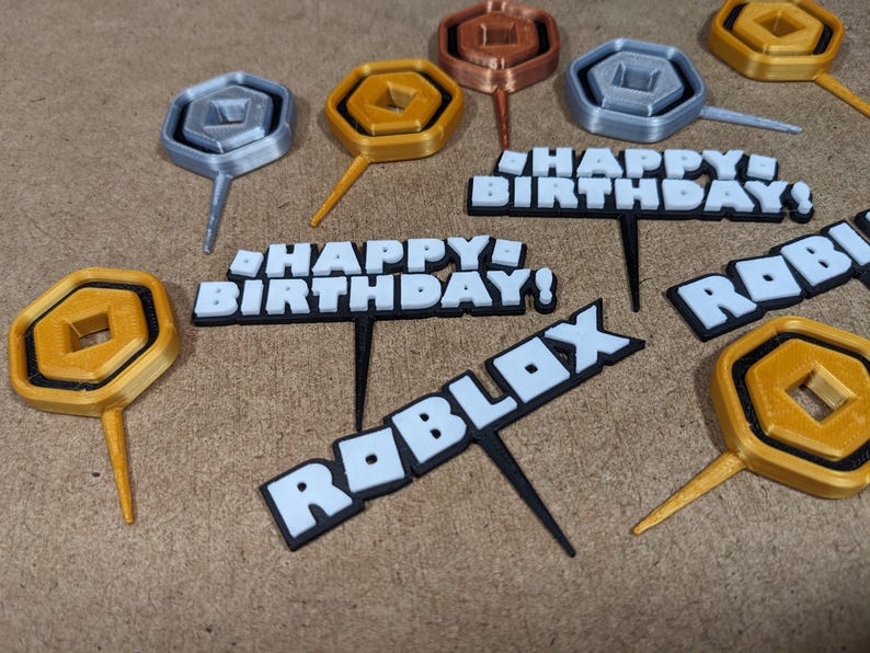 Op de afbeelding: Een set van 3D-geprinte taarttoppers met een Roblox-thema. De toppers bevatten een gouden munt, een zilveren munt en de woorden "Happy Birthday!" en "Roblox".