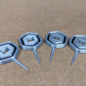 Op de afbeelding: Vier zilverkleurige, 3D-geprinte, zeshoekige gereedschappen met een klein vierkant gat in het midden. Elk gereedschap heeft een puntige punt.