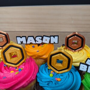 Op de afbeelding: Een close-up van cupcakes versierd met gouden en koperen Roblox-gamemunten en een witte topper met de tekst "MASON".