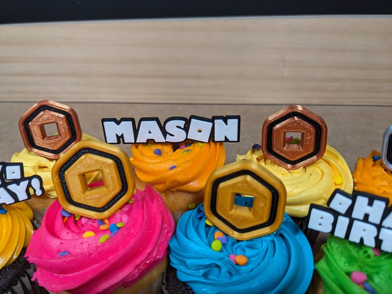 Op de afbeelding: Een close-up van cupcakes versierd met gouden en koperen Roblox-gamemunten en een witte topper met de tekst "MASON".