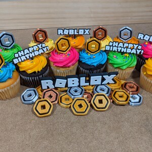 Op de afbeelding: Een set van zes cupcakes versierd met kleurrijk glazuur en Roblox-thema toppers. De cupcakes zijn in een cirkel op een bruin oppervlak gerangschikt. De toppers bevatten de woorden "Roblox" en "Happy Birthday!" en zilveren en gouden munten.