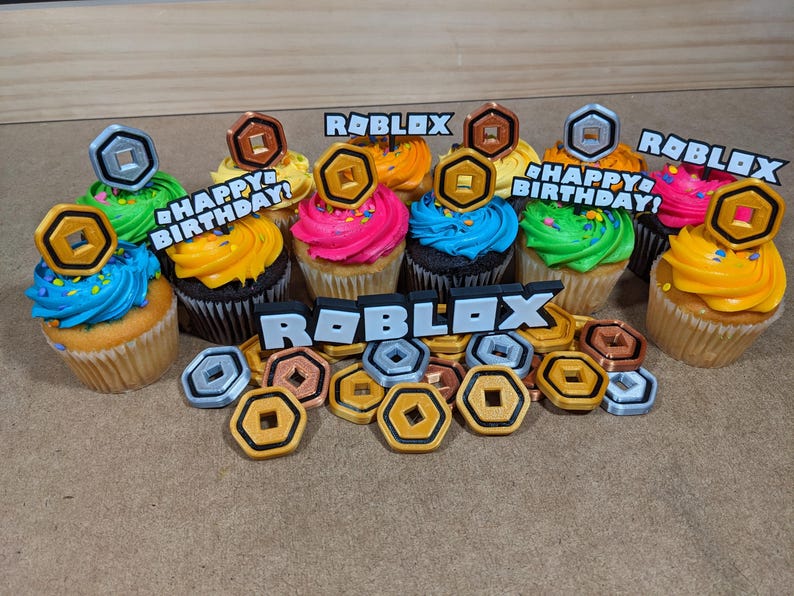 Op de afbeelding: Een set van zes cupcakes versierd met kleurrijk glazuur en Roblox-thema toppers. De cupcakes zijn in een cirkel op een bruin oppervlak gerangschikt. De toppers bevatten de woorden "Roblox" en "Happy Birthday!" en zilveren en gouden munten.