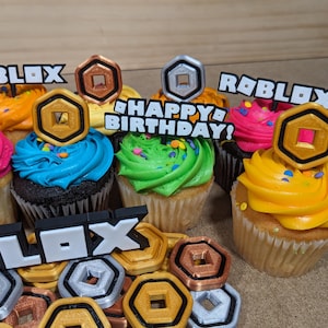 Op de afbeelding: Zes cupcakes versierd met kleurrijk glazuur en sprinkles. Elke cupcake heeft een andere Roblox-thema topper, waaronder een gouden munt, een zilveren munt en de woorden "Roblox" en "Happy Birthday!"