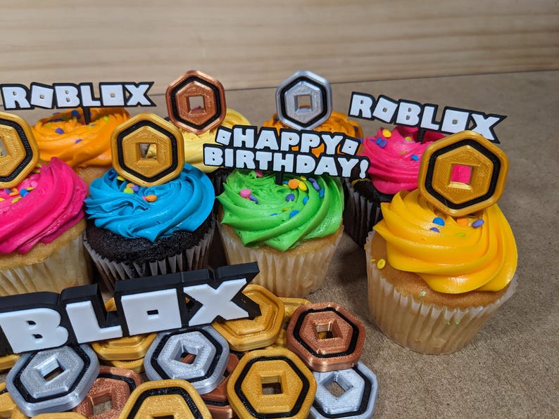 Op de afbeelding: Zes cupcakes versierd met kleurrijk glazuur en sprinkles. Elke cupcake heeft een andere Roblox-thema topper, waaronder een gouden munt, een zilveren munt en de woorden "Roblox" en "Happy Birthday!"