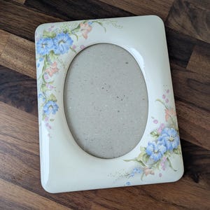 May include: Cream-colored oval picture frame with floral accents. The frame features a rectangular shape with rounded corners and an oval opening. Blue, pink, and green floral designs adorn the corners, adding a touch of elegance.