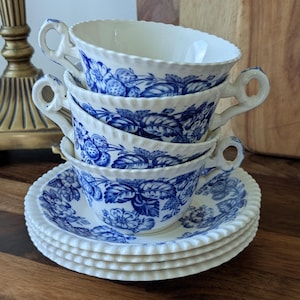 May include: A stack of three white ceramic teacups and three matching saucers, each featuring a blue floral and strawberry pattern. The cups have scalloped rims and looped handles, while the saucers have a matching scalloped, rope-like edge.