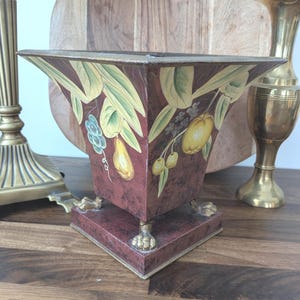 May include: A burgundy-colored, square vase with a flared top, featuring hand-painted fruit and leaf designs. The vase rests on a square base with gold-colored claw feet. A decorative piece for home decor.