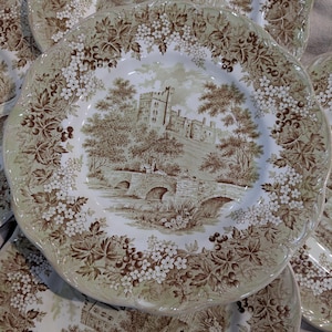 May include: A collection of vintage ceramic plates with a brown and white transferware design. Each plate features a central scene of a castle and bridge, surrounded by floral and berry motifs. The plates have a scalloped edge.
