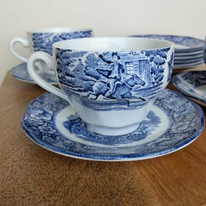 May include: A set of blue and white ceramic teacups and saucers. The cups and saucers feature a detailed pastoral scene. The cups have a curved handle and sit on matching saucers. Additional plates are stacked in the background.