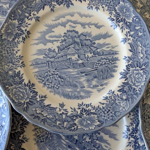 May include: A collection of blue and white ceramic plates. Each plate features a central landscape scene with a bridge and house, surrounded by a floral border. The plates have a scalloped edge and are arranged in a slightly overlapping pattern.