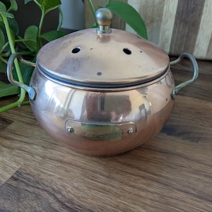 May include: A round copper potpourri container with a domed lid featuring small ventilation holes and a central knob. The vessel has two curved metal handles on opposite sides and a rectangular brass plaque on the front with the word POTPOURRI.
