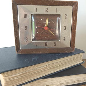 May include: Vintage General Electric alarm clock with a brown wooden frame and a silver face. The clock has a dark brown clock face with red and gold hands. The word "ALARM" is visible on the clock face. The clock is sitting on top of two blue books.