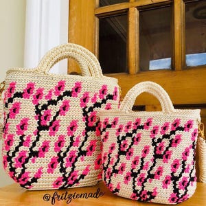 Könnte beinhalten: Zwei gehäkelte Tragetaschen in Creme mit rosa und schwarzen Blumenmustern. Die Taschen haben cremefarbene Griffe und Verzierungen. Die Taschen stehen auf einer Holzoberfläche. Der Text "@fritziemade" ist sichtbar.
