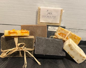 May include: Assortment of handmade goat milk soap bars. The bars are wrapped in brown paper and tied with twine. The soap bars are in various colors and scents, including charcoal, brown, and orange. One bar has the text "Goat Milk Soap Hand Poured by Mel" on the label.