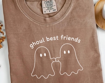 May include: A brown short-sleeved t-shirt with a crew neckline featuring a white line-art graphic of two ghosts holding hands. The text "ghoul best friends" is printed in white above the ghosts. A Comfort Colors brand tag is at the neck.