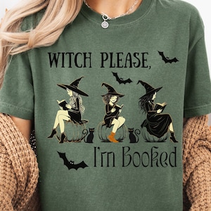 May include: Green t-shirt with the text "Witch Please, I'm Booked." The shirt features a graphic of three witches reading books, sitting on pumpkins, with bats and black cats. The shirt is a comfortable, casual style.