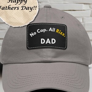 May include: A gray baseball cap with a curved brim. A black patch on the front reads "No Cap. All Rizz. DAD" in white and yellow text. A circular graphic above the cap says "Happy Fathers Day!!"