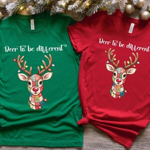 May include: Green and red t-shirts with a reindeer graphic and the text "Deer to be different." The reindeer has a colorful patchwork design, antlers with lights, and a red nose. Festive holiday apparel.