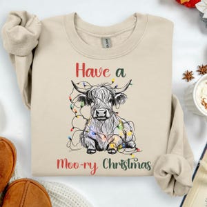 May include: A beige sweatshirt with a graphic of a Highland cow decorated with Christmas lights. The text "Have a Moo-ry Christmas" is printed in red and green. The sweatshirt has a crew neck and long sleeves.
