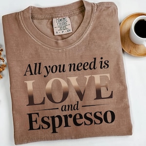 May include: A brown Comfort Colors t-shirt with the phrase "All you need is LOVE and Espresso" printed in a gradient and black font. A small tag is visible at the neckline. A cup of coffee sits on a wooden coaster in the background.