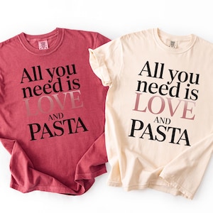 May include: Two t-shirts, one red and one cream-colored, with the phrase "All you need is LOVE AND PASTA" printed in black and pink lettering. The shirts are casual and likely made of cotton.