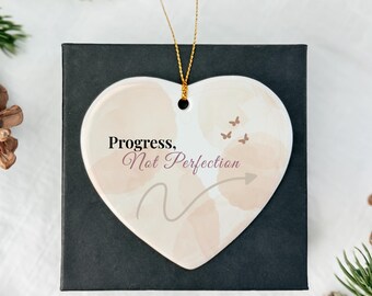 May include: Heart-shaped ceramic ornament with the text "Progress, Not Perfection" in a script font. The ornament has a watercolor design in shades of beige and peach, with three small butterfly illustrations and a gray arrow. A gold string is attached for hanging.