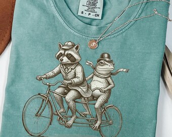 May include: Teal-colored t-shirt featuring a raccoon and frog riding a tandem bicycle. The raccoon wears a suit and hat, while the frog sports a scarf and bowler hat. The text "Get on loser, we're writing IEPs" is printed at the bottom.