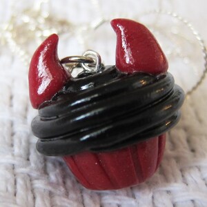 May include: A red and black cupcake charm with devil horns. The cupcake is decorated with black frosting and red sprinkles. The charm is attached to a silver chain.