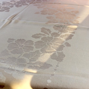 May include: Cream-colored tablecloth with a floral pattern. The design features large, stylized flowers and leaves in a lighter shade of cream, creating a subtle embossed effect. The fabric appears to be a soft, woven material, suitable for dining or special occasions.