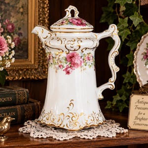May include: A white porcelain coffee pot featuring a floral pattern of pink roses and green leaves, accented with intricate gold scrollwork along the base, handle, and rim. The pot includes a matching lid with a gold-trimmed handle and spout.