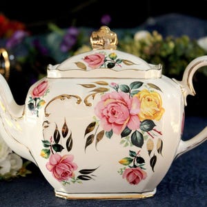 May include: Cream-colored porcelain teapot with a gold-colored crown-shaped finial and gold trim. The teapot is decorated with pink and yellow roses, green leaves, and gold accents. The handle and spout are curved.