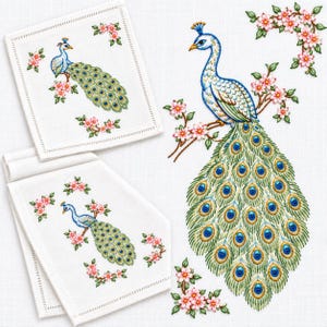 May include: Two white fabric napkins featuring an embroidered peacock perched on a flowering branch with pink blossoms. The peacock has a long, detailed tail with blue and gold eye-shaped patterns. Additional pink floral clusters surround the bird.