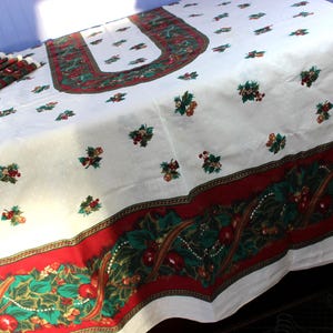 May include: A white Christmas tablecloth with a red and green border featuring holly and berry designs. The tablecloth has a repeating pattern of holly and berries scattered across the surface. Matching napkins are visible.