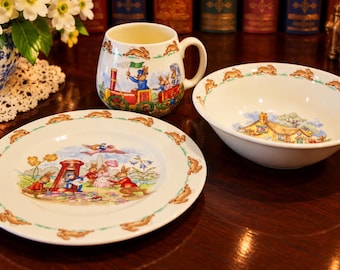 May include: A ceramic three-piece dinnerware set featuring a plate, a bowl, and a mug. Each piece is decorated with illustrations of rabbits, a red telephone booth, a train, and a cottage, with a repeating rabbit border pattern.