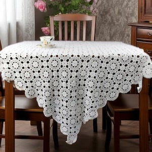 May include: A white, crocheted tablecloth with a circular pattern covers a wooden table. The tablecloth has a scalloped edge and is draped over the table. A teacup and saucer sit on the table. The table is surrounded by wooden chairs.
