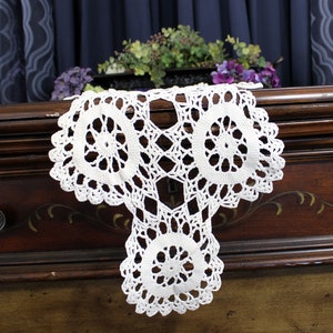 May include: White crocheted doily with three circular designs, draped over a dark brown wooden dresser with a drawer.