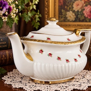 May include: A white porcelain teapot featuring a delicate pattern of small red roses with green leaves. The teapot has a gold-trimmed lid, spout, handle, and base, with a ribbed texture along the lower body and a gold embossed rim on the lid.
